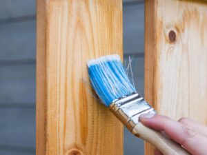 A hand painting a wooden fence plank with a blue paintbrush outdoors. Perfect for DIY and renovation themes.